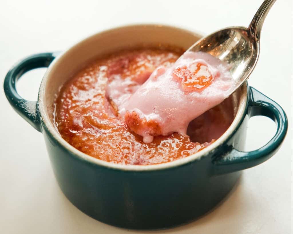 A spoon scoops up pink, creamy Strawberry Crème Brûlée from a small, round, blue baking dish with handles, revealing a smooth golden-brown top layer and a soft, airy interior—an easy recipe for an elegant dessert.
